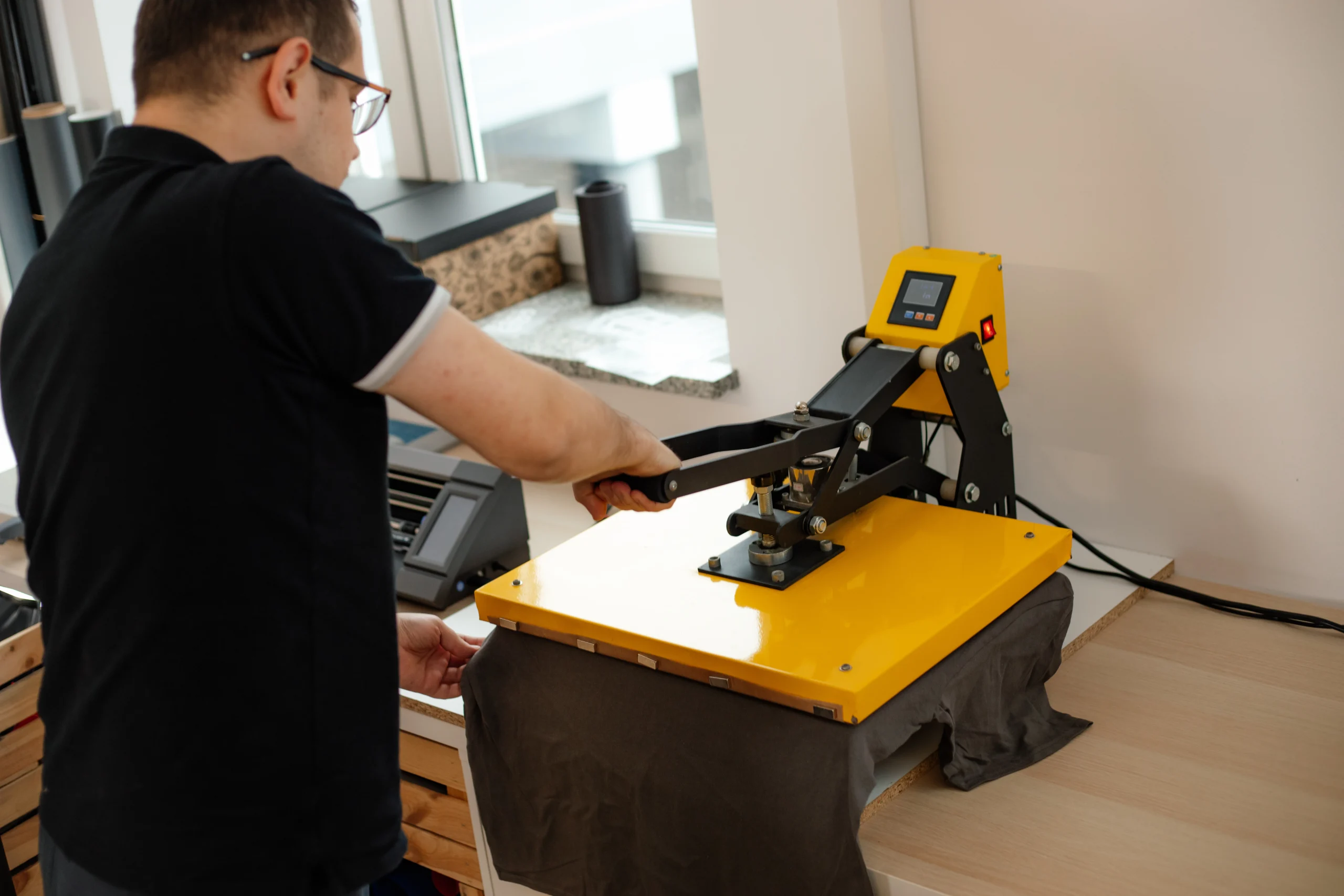 VistaCreate-548641908-stock-photo-man-worker-glasses-back-using-heat-thermal-textile-application-transfer-machine-printing-application-t-shirt
